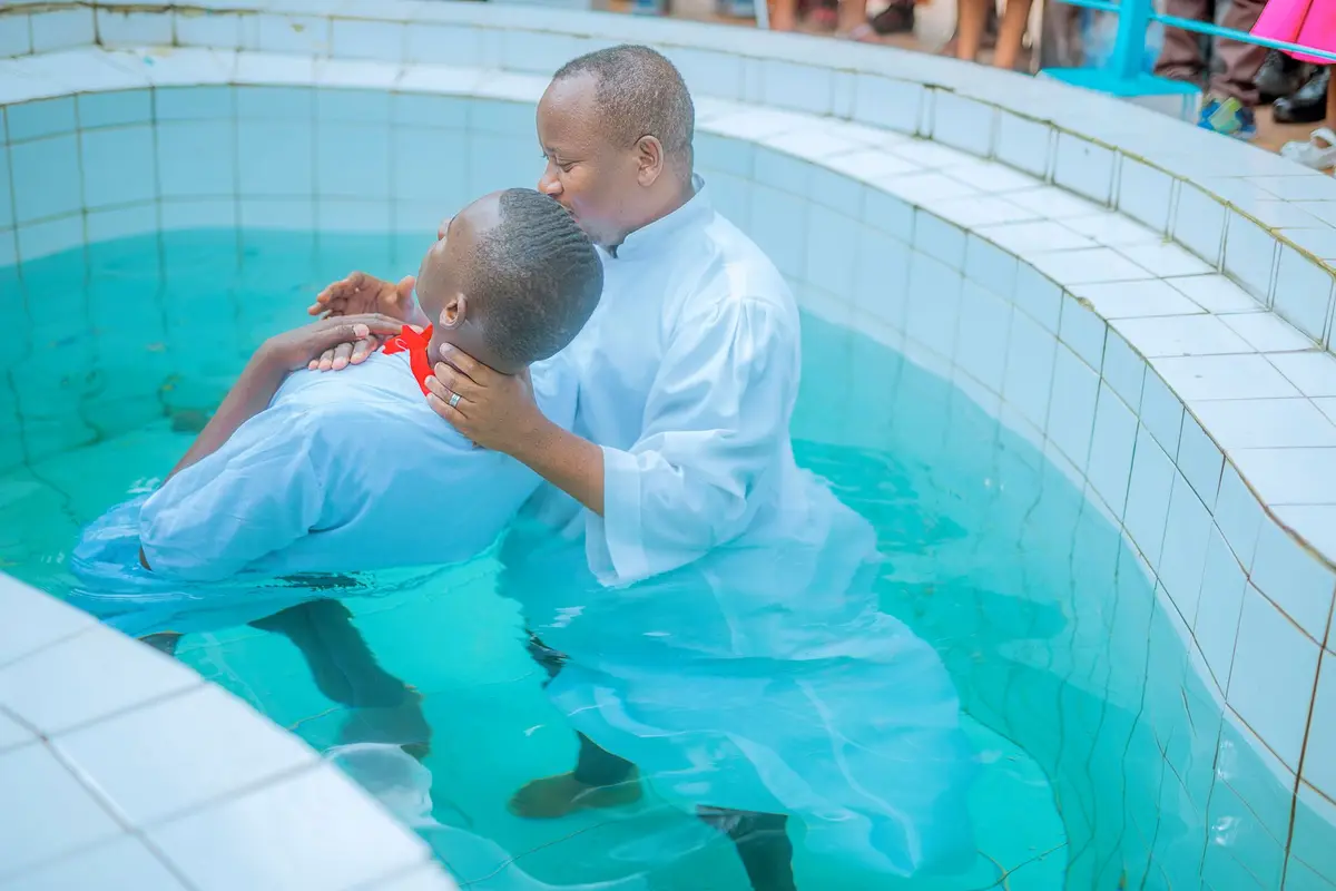 A pastor in white baptizing a man in a white robe in a turquoise baptism pool