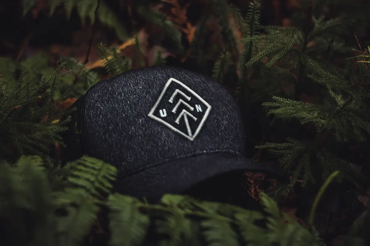 A dark heathered cap with a white embroidered geometric logo among green ferns