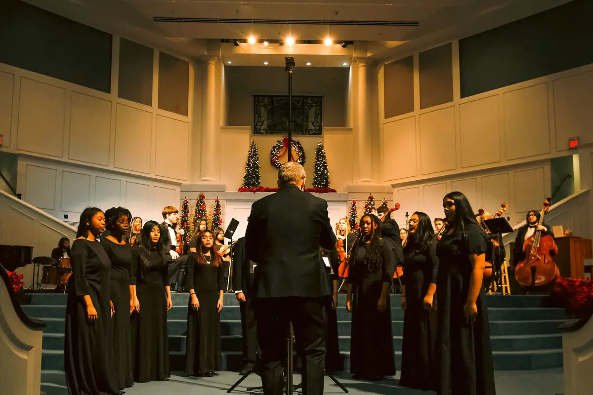 A choir in black singing at a church decorated for Christmas while a director conducts