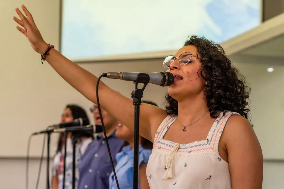 A woman with her hand raised singing into a microphone with a church choir behind her