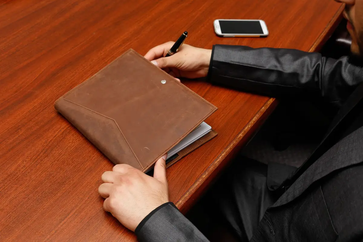 A person in a dark suit writing in a brown leather portfolio at a wooden desk