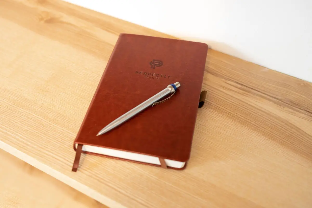 A brown leather notebook with a debossed logo and a silver pen on a light wood table