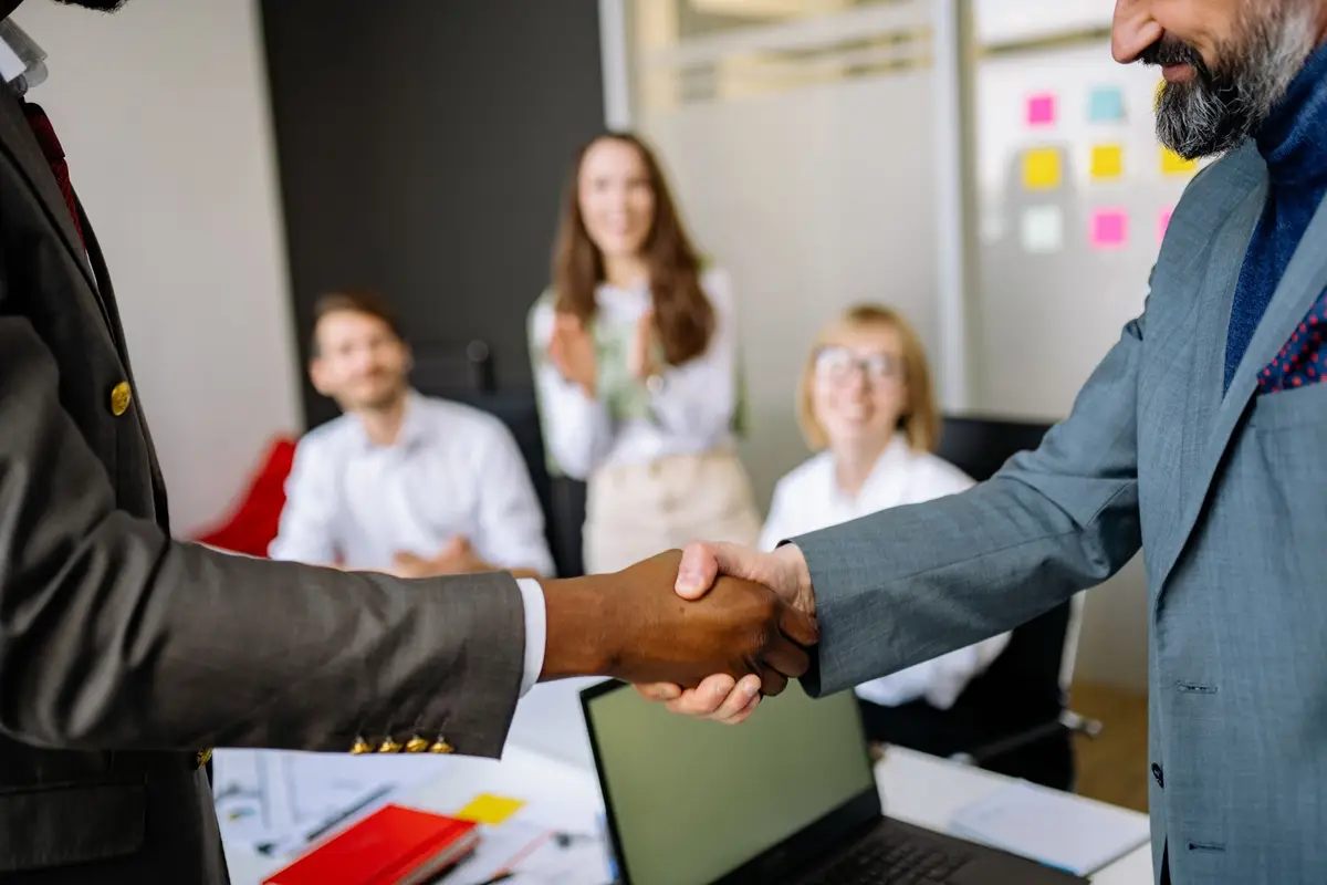 Two businessmen shaking hands in an office while colleagues smile and clap in the background