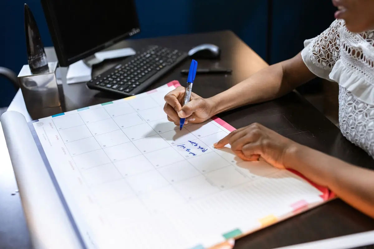 A person writing notes on a large desk calendar with a blue pen