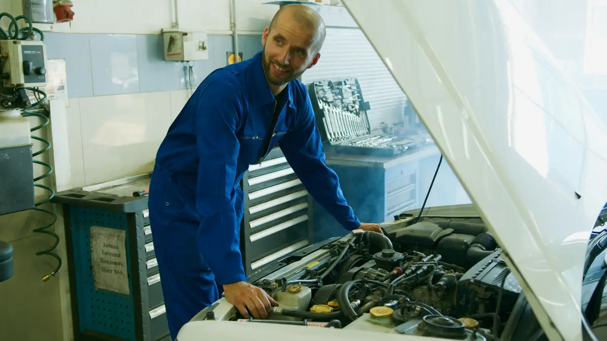 A smiling mechanic in blue coveralls working on a car engine in a shop