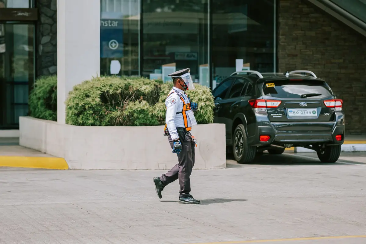 A security guard in uniform patrolling past a parked car outside a building