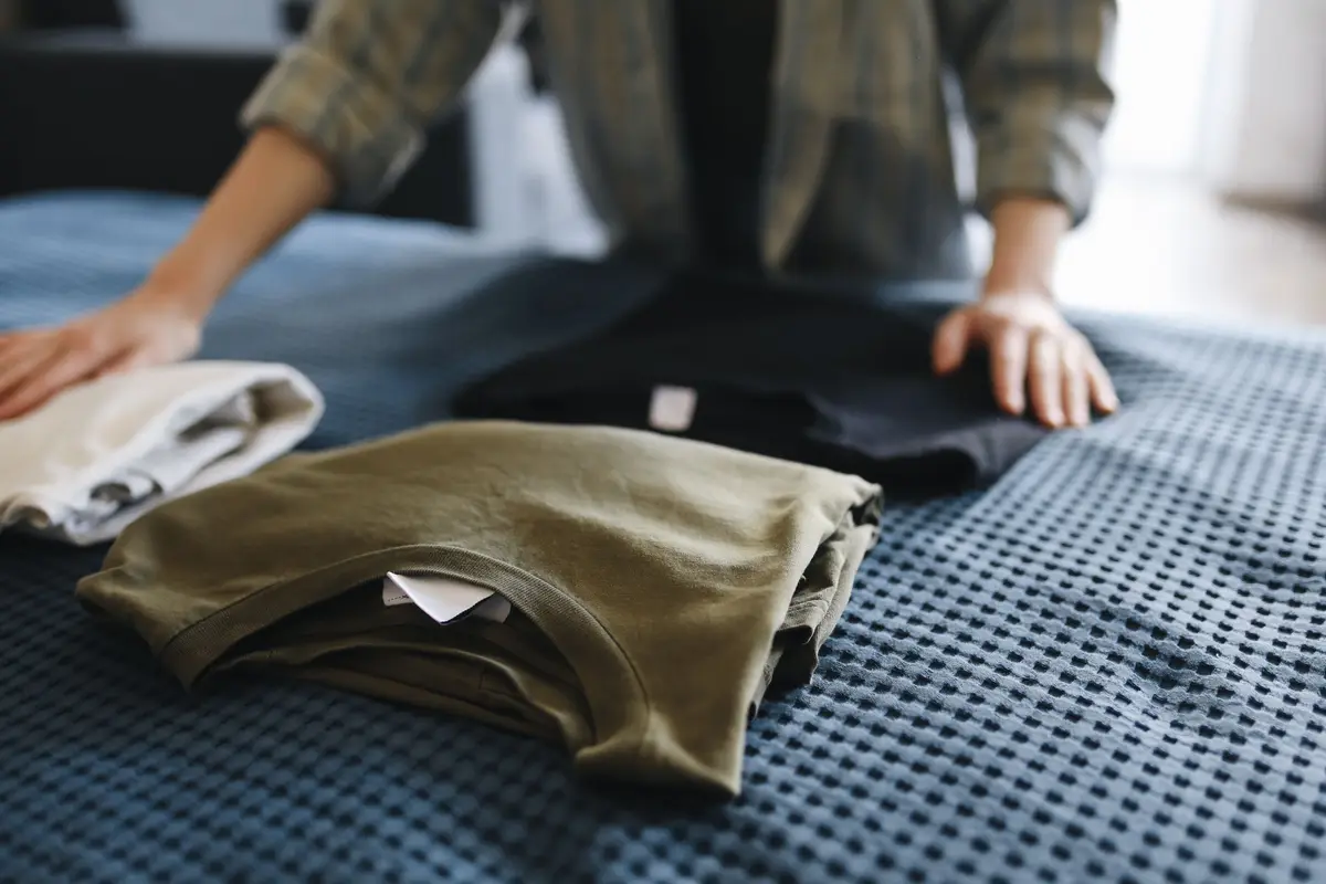 Hands sorting and folding a stack of t-shirts on a bed