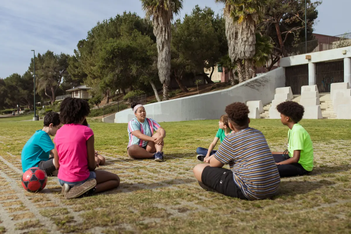 A small group of kids sitting in a circle on the grass with a leader
