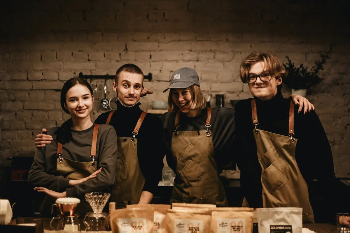 Four cheerful coffee shop employees in matching aprons behind the counter