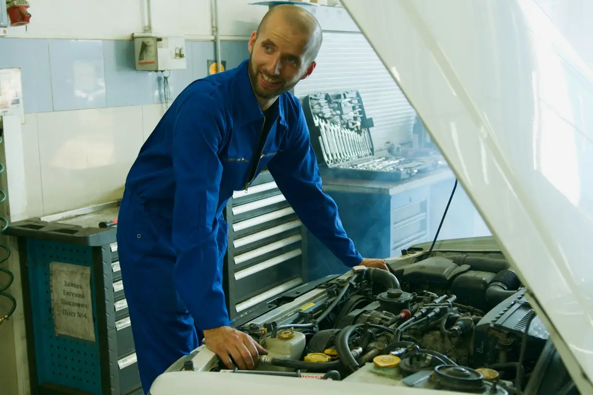 A smiling mechanic in blue coveralls working on an engine in a garage