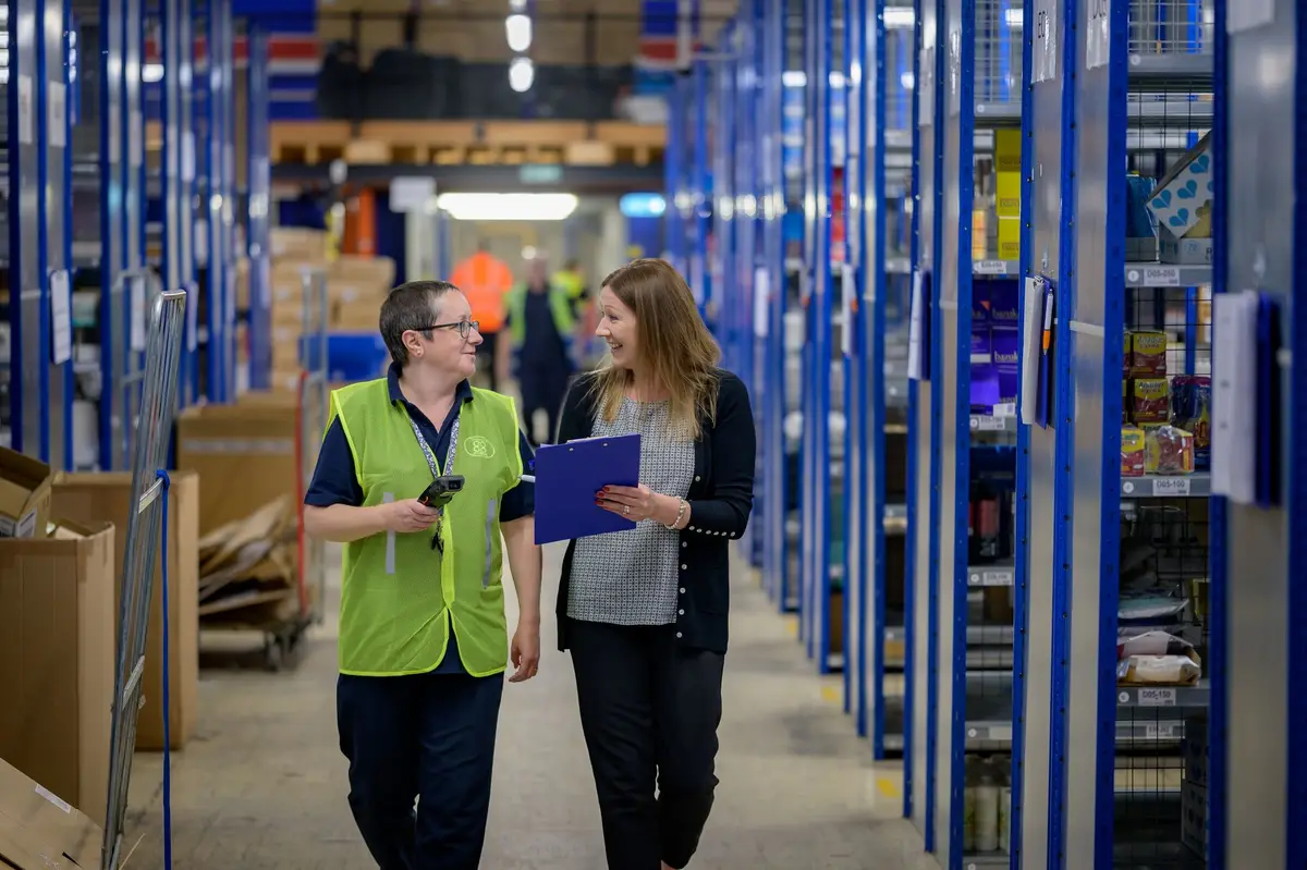 A warehouse worker in a high-visibility vest talking with a colleague in a stockroom aisle