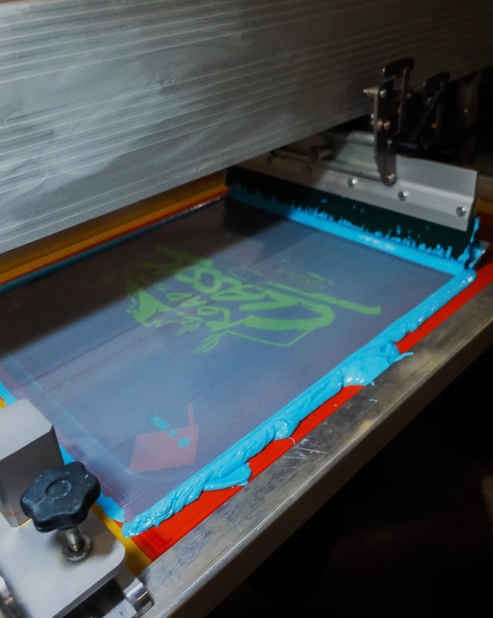 A screen printing screen loaded with cyan ink and a squeegee on the press at Heart & Hook